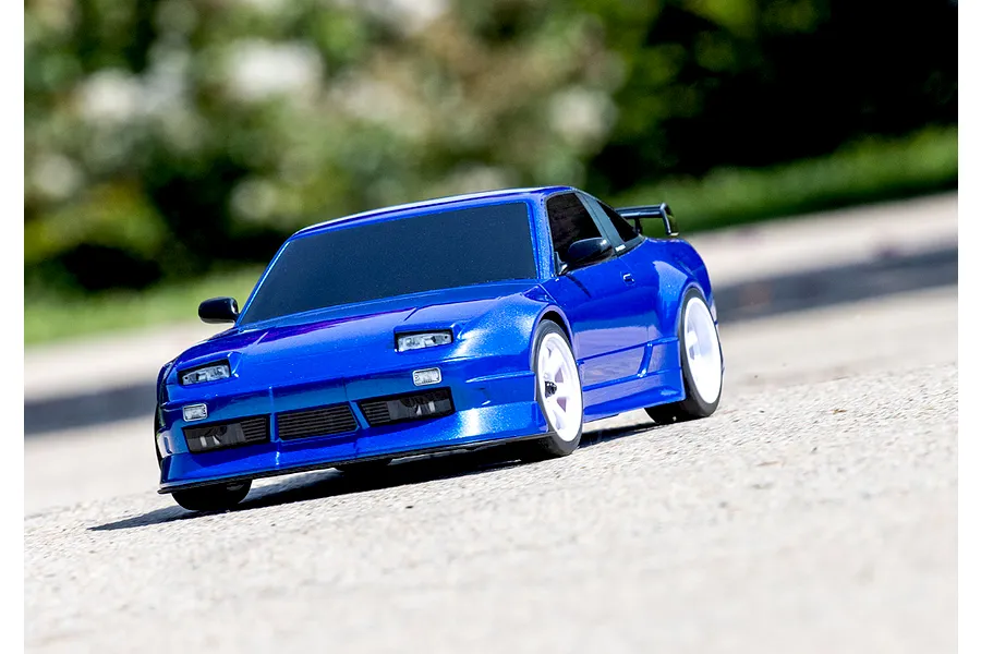 A blue Traxxas Nissan 240SX RC drift car driving on a paved surface with a blurred outdoor background.