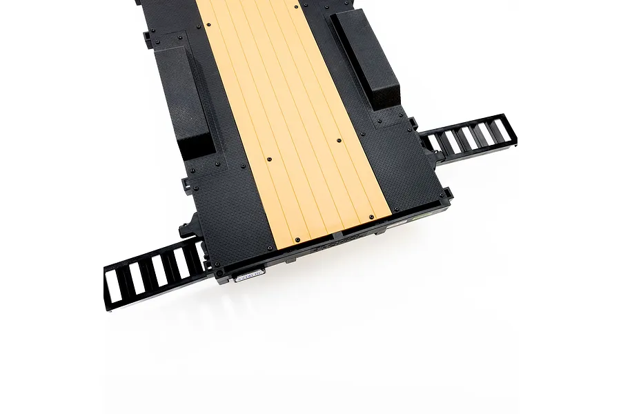 A top-down view of an Axial 1/10 scale flat bed RC vehicle trailer featuring a black diamond-plate deck with a tan wood-look center section and two fold-out ramps.