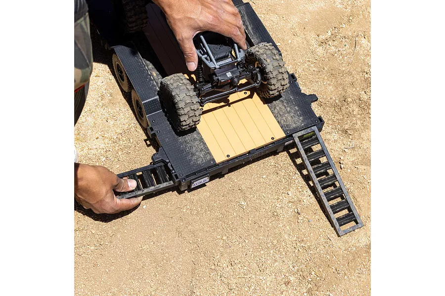 A person's hands positioning a small RC crawler onto an Axial 1/10 scale flatbed trailer with deployed loading ramps on a dirt surface.