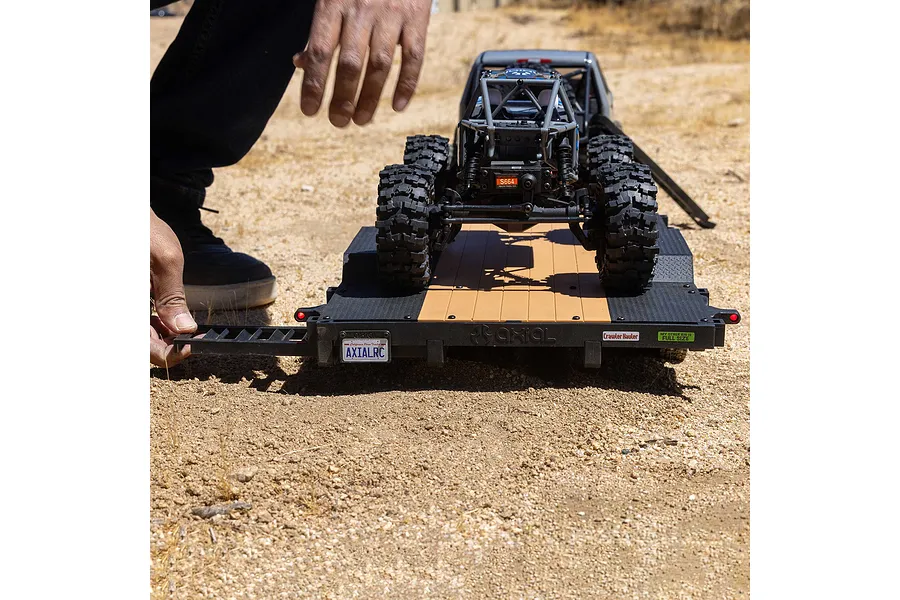 A person's hand adjusting the ramp on an Axial 1/10 scale flat bed vehicle trailer with an RC crawler loaded on top, sitting on a dirt surface.