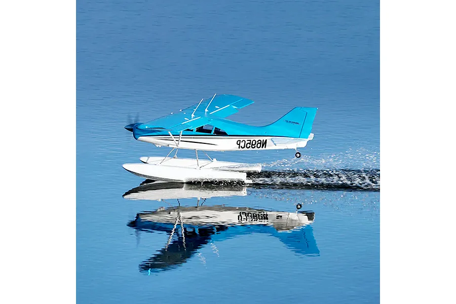 A blue and white FMS Maule RC plane with floats taking off from the surface of calm blue water, creating a wake and reflection.