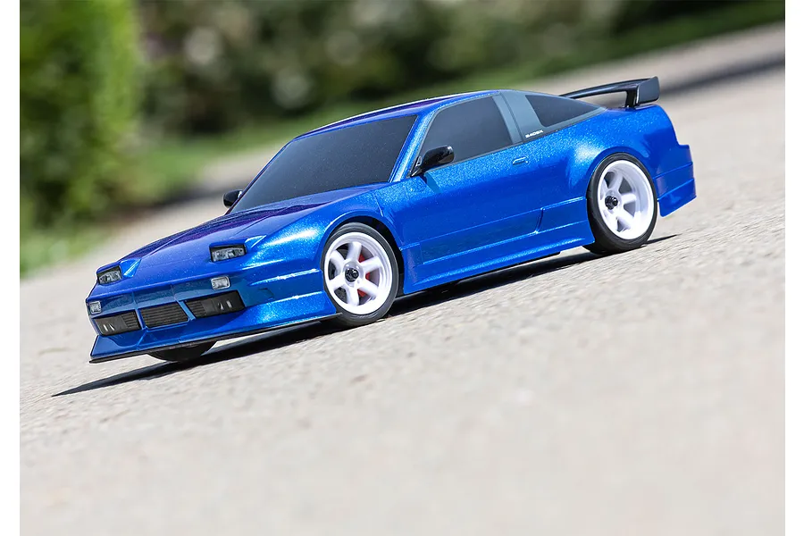 A blue Traxxas Nissan 240SX RC drift car on a paved surface with white wheels and a rear spoiler.