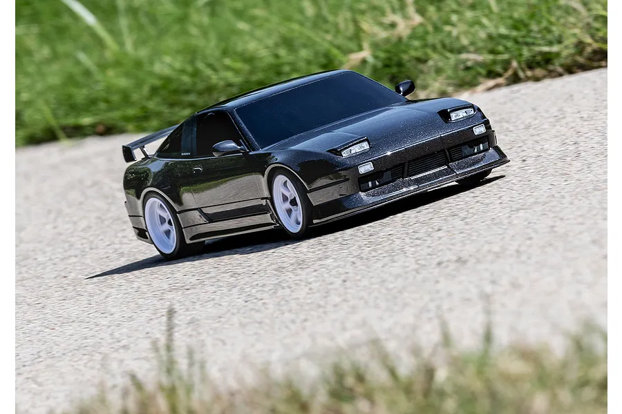 A black Traxxas Nissan 240SX RC drift car driving on a paved surface with grass in the background.
