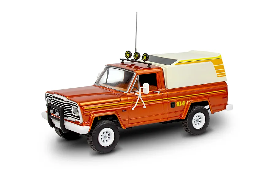 A 1/24 scale model of an orange Jeep J-10 pickup truck with a white camper shell, roof lights, and off-road tires, shown against a white background.