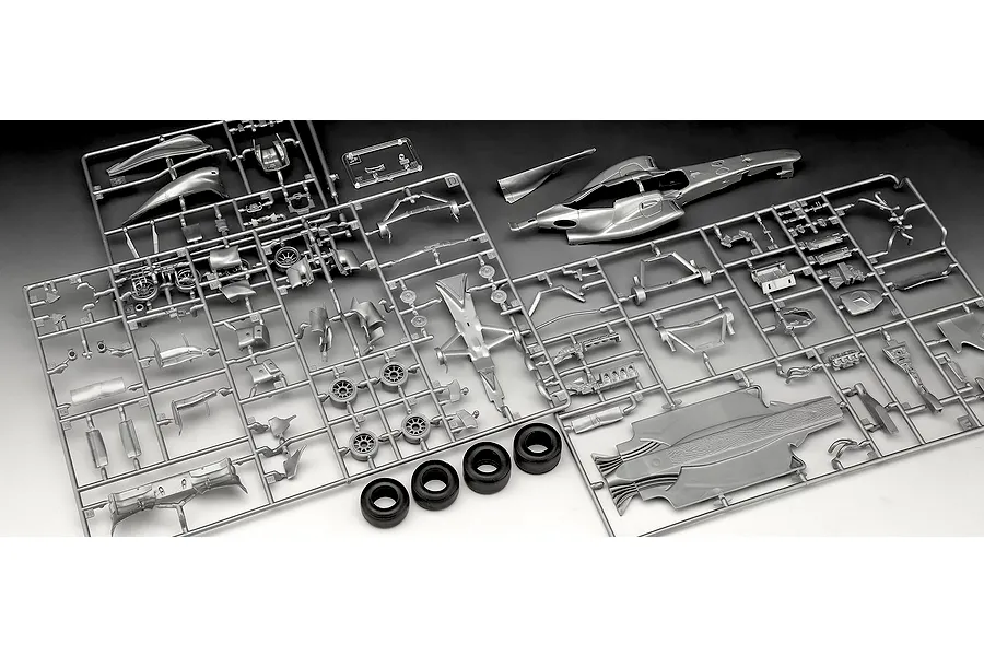 Silver plastic model kit sprues for a Formula 1 race car, including body panels, wheels, and chassis parts, laid out on a white surface.