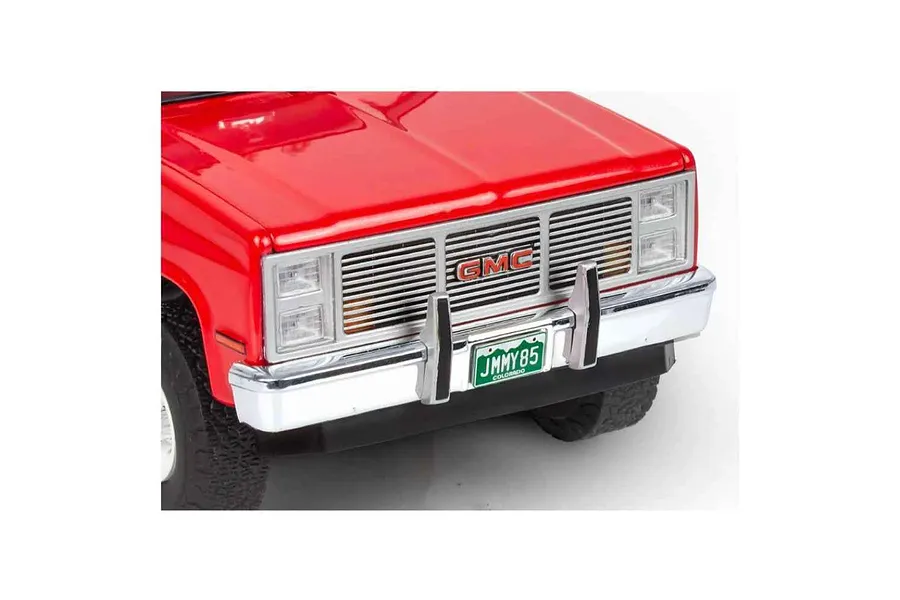 Close-up of the front end of a red 1985 GMC Jimmy model truck featuring a chrome grille, bumper, and a Colorado license plate reading JIMMY85.