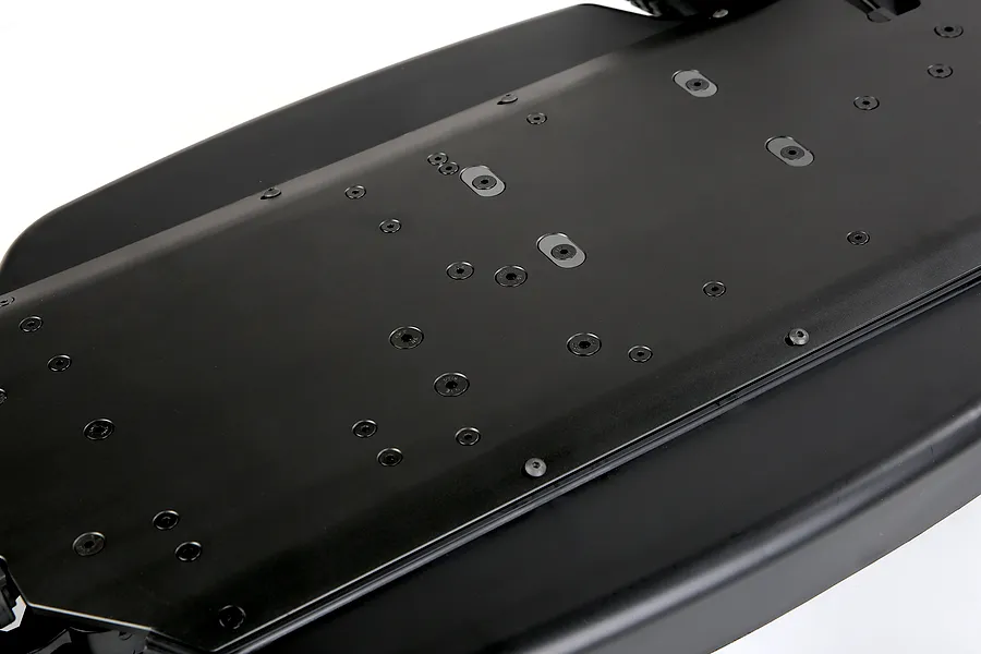 A close-up, angled view of a black aluminium RC car main chassis showing the underside with numerous countersunk screw holes.