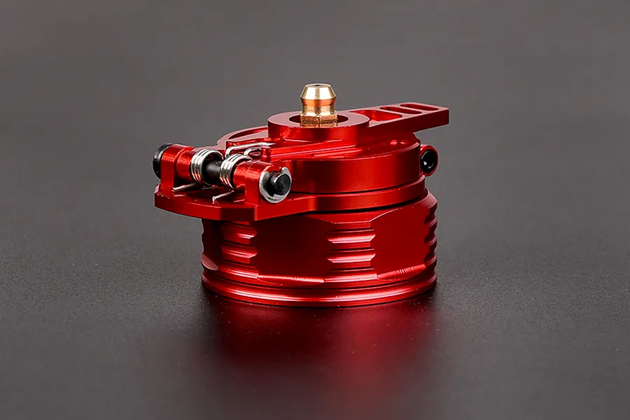 A red anodized aluminium RC fuel tank cap with a spring-loaded hinge and brass breather nipple, set against a dark grey background.