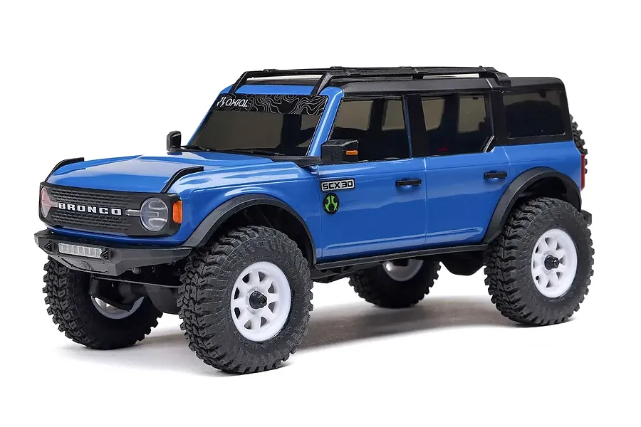 Blue Ford Bronco RC rock crawler studio product shot against a clean white background, showcasing its black roof rack, white wheels, and aggressive tread tires.