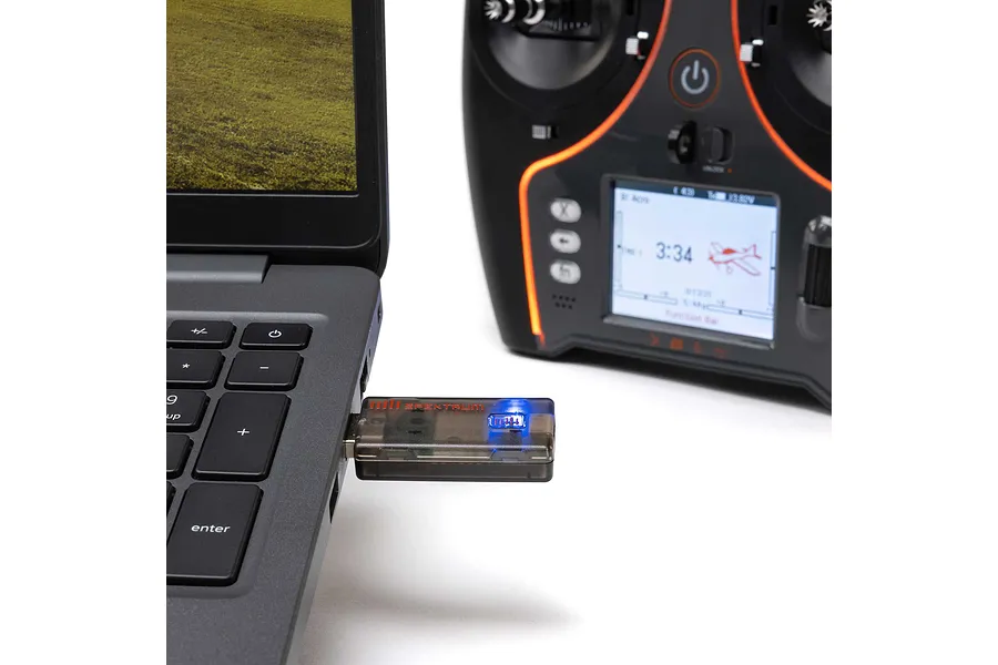 Spektrum wireless simulator USB dongle plugged into a laptop alongside an RC transmitter on a white surface.