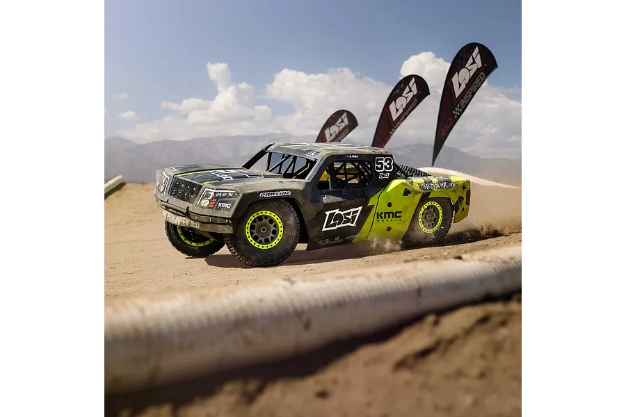 Losi 1/5 scale short course truck driving fast on a dirt track under a sunny sky with flags in the background.