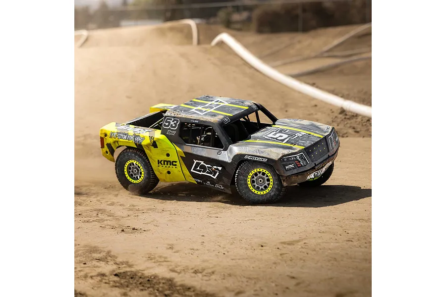 Losi 1/5 scale short course RC truck driving on a dirt track, featuring yellow and black livery with mud splatters.