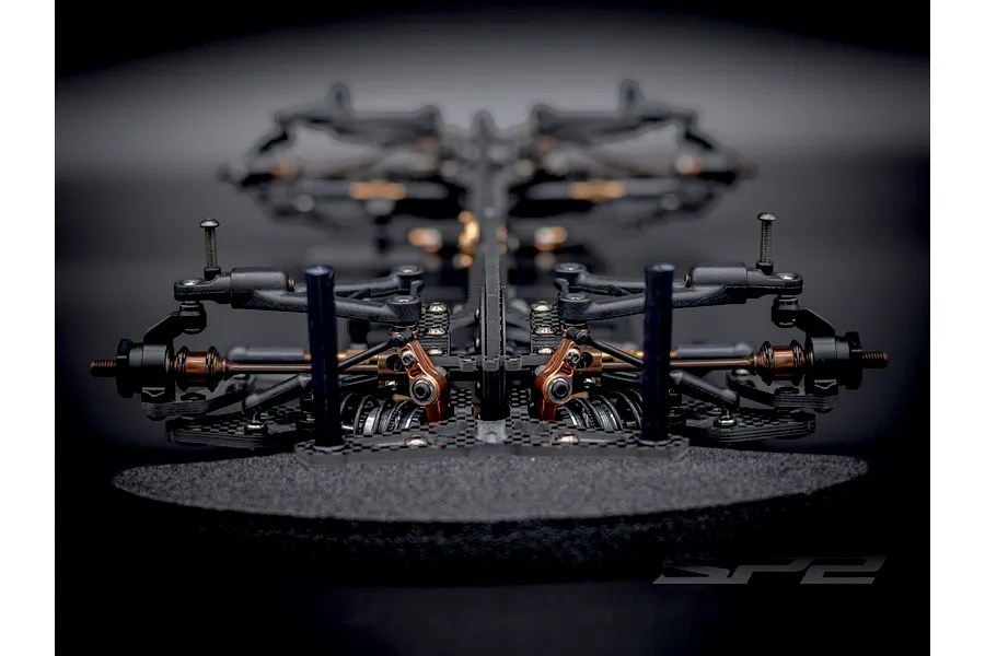 Low angle front view of the RC Maker SP2 EP touring car chassis showing carbon fiber, metal, and plastic components on a dark background.