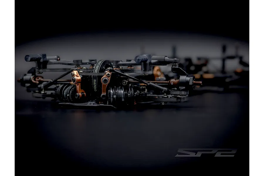Close-up studio shot of the RC Maker SP2 EP touring car front suspension, showing black and bronze components and drive belts on a dark background.