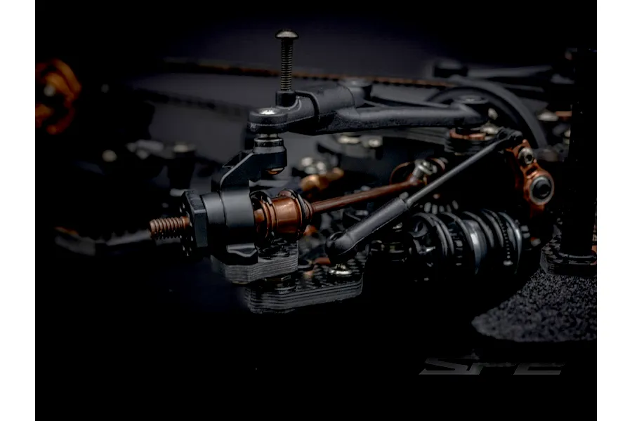 Close-up studio shot of the RC Maker SP2 EP RC touring car front suspension, showing carbon fiber, black plastic, and bronze-colored metallic parts.