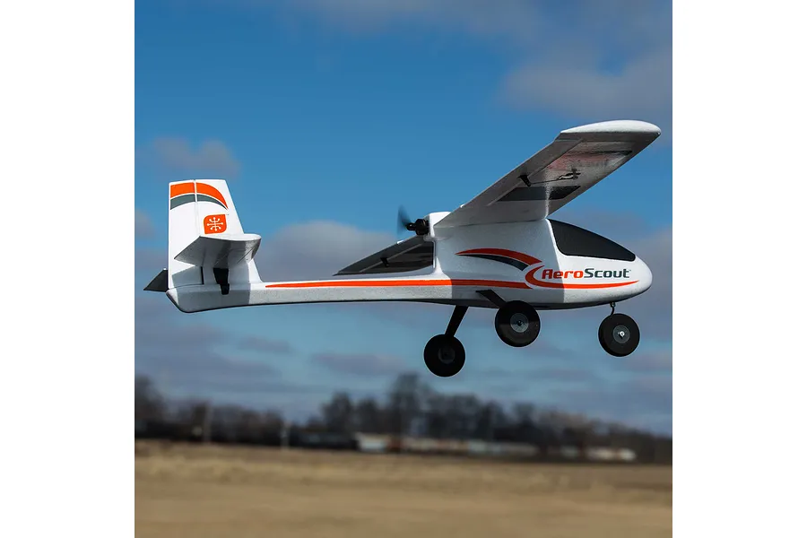 HobbyZone AeroScout S 2 RC airplane flying outdoors against a blue sky with clouds and a distant horizon.