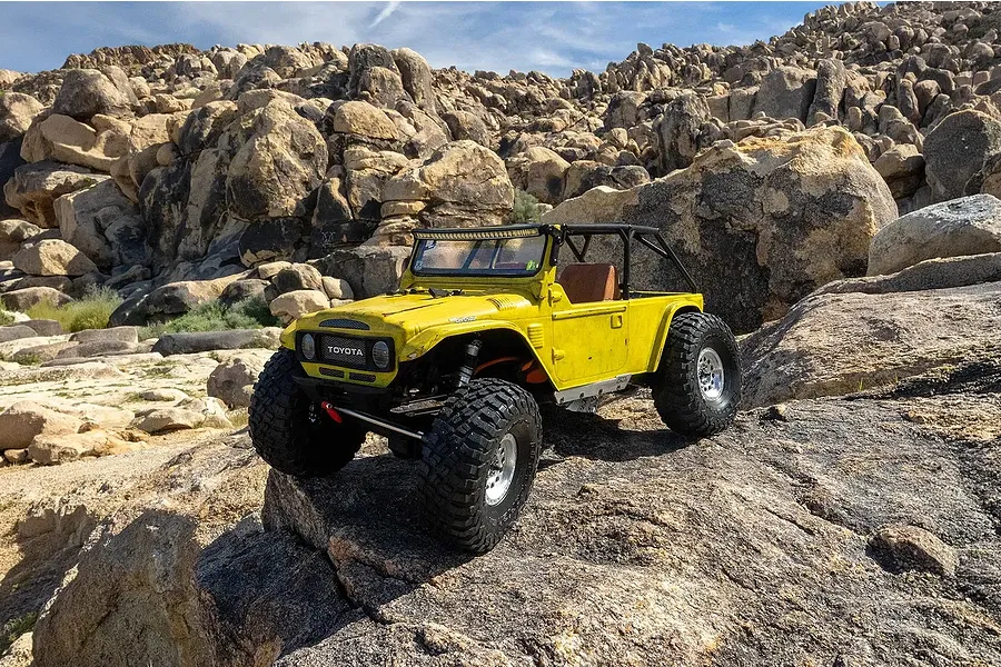 A yellow RC rock crawler with a roll cage traversing a rocky outdoor landscape in daylight.