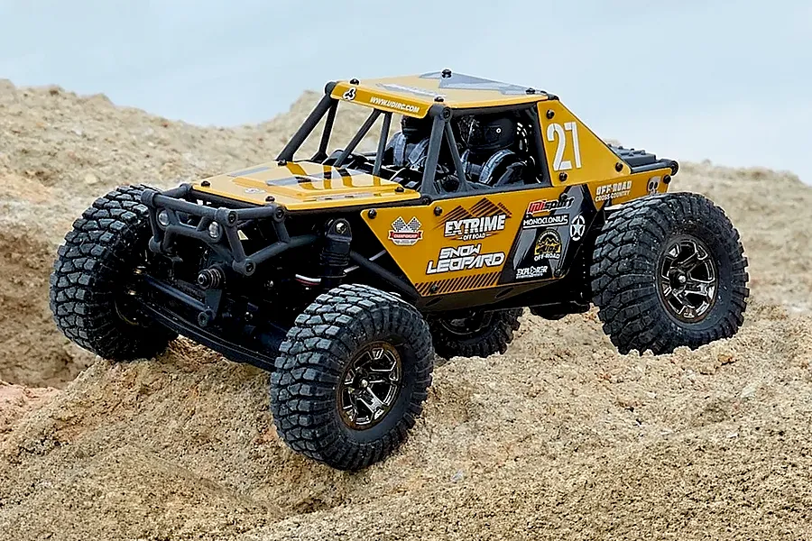 UDI UCX24 Snow Leopard 1/24 scale yellow RC rock crawler parked on sandy terrain outdoors.