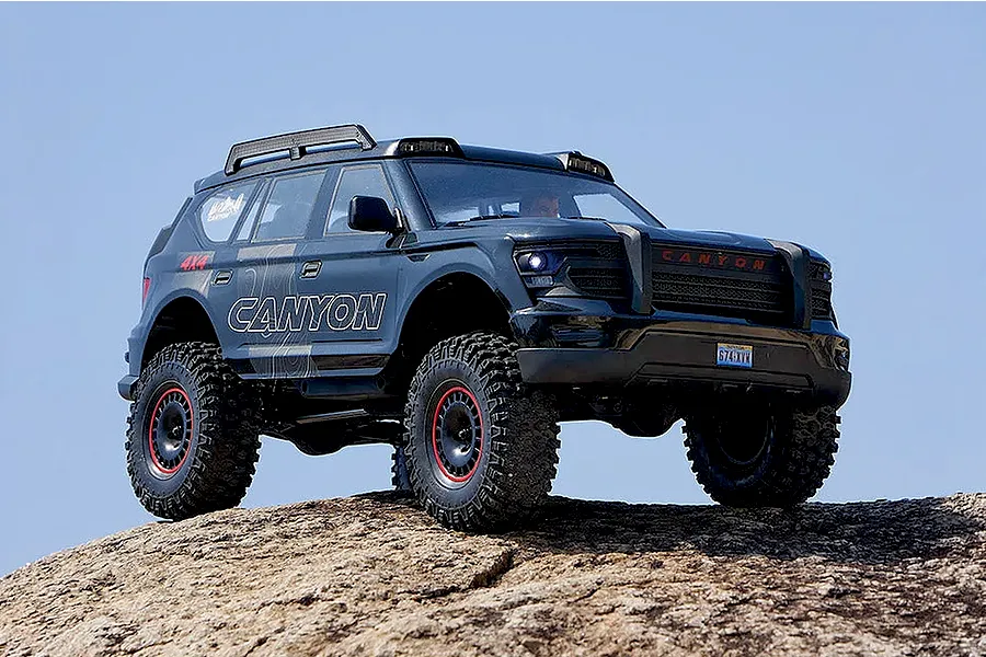 FMS Canyon RS RC rock crawler driving on a rocky outdoor surface against a clear blue sky background.