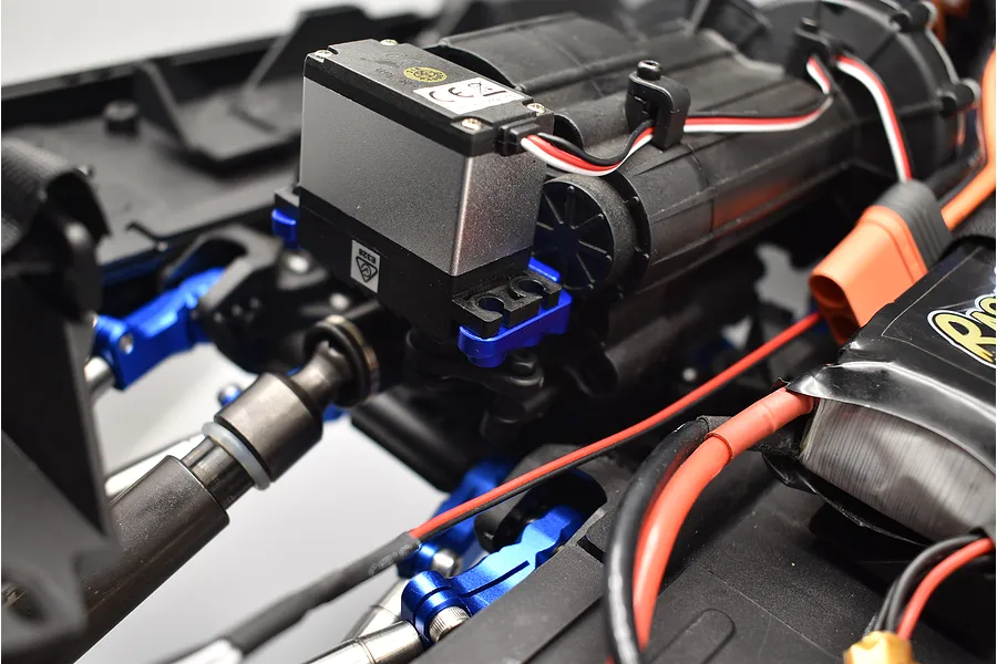 Close-up of an RC car chassis showing a blue aluminium 2-speed transmission servo mount installed near the transmission and servo.