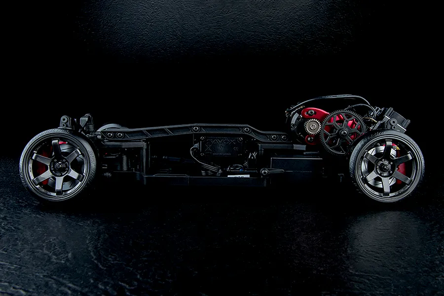 Side profile of an MST RC drift car chassis showing electronics, gears, and dark grey wheels against a black background.
