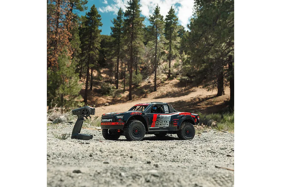 ARRMA Mojave 6S EXB desert truck and remote control parked on a dirt path surrounded by pine trees outdoors.