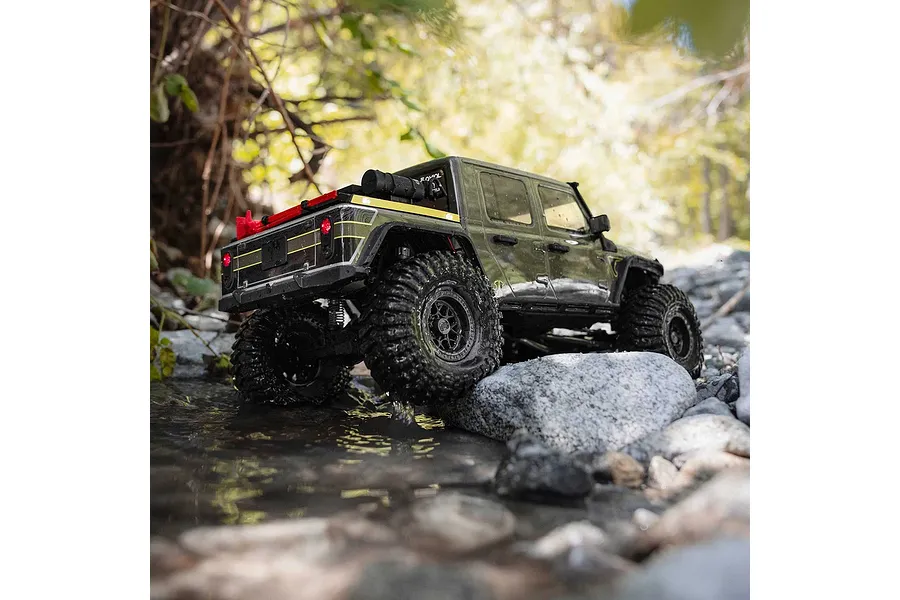 Green Axial RC rock crawler truck driving through a shallow rocky stream outdoors.