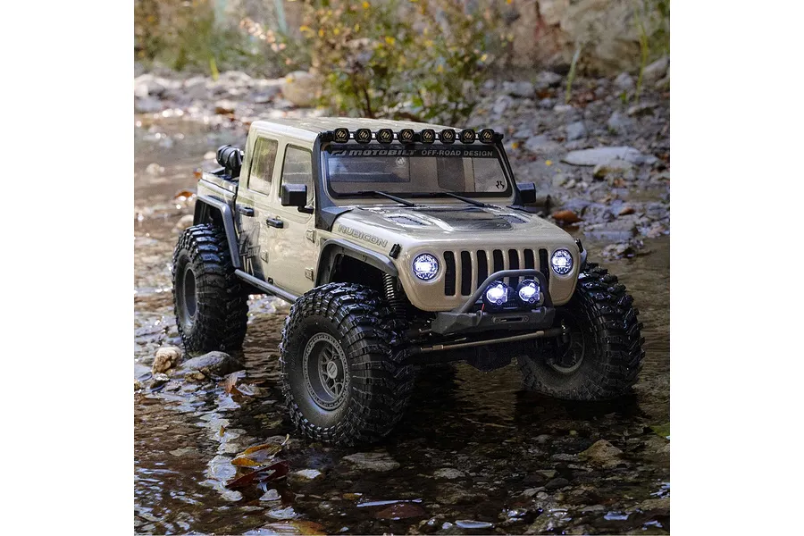 Tan Axial SCX10 III RC rock crawler driving through a shallow rocky creek with active headlights