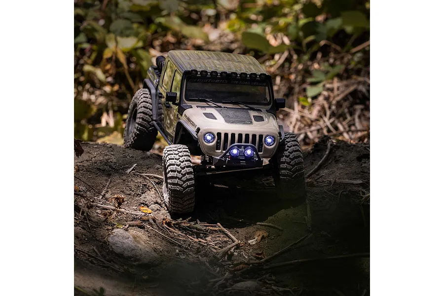 Tan Axial RC rock crawler truck navigating an outdoor dirt trail with illuminated headlights and light bar.