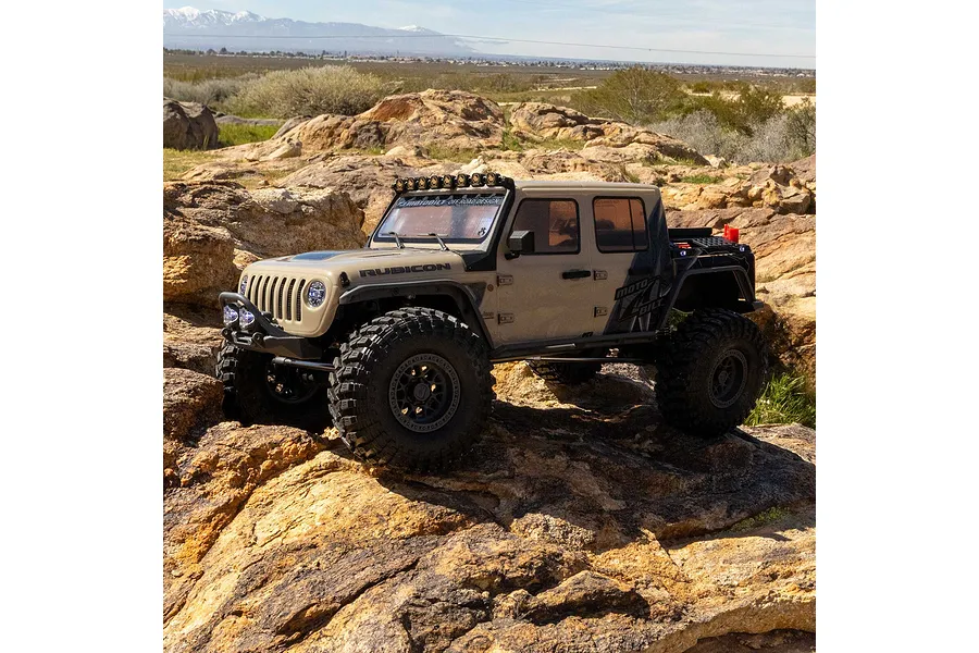 Tan Axial SCX10 III Motobilt Jeep Gladiator RC crawler driving over rocky outdoor terrain.