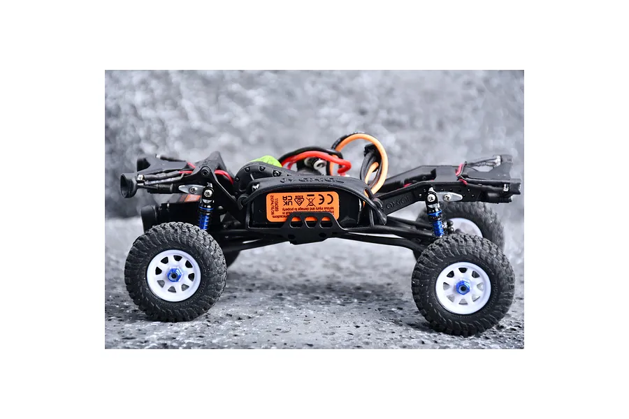 Side view of a small RC crawler chassis featuring blue suspension shocks and white wheels on a textured surface.