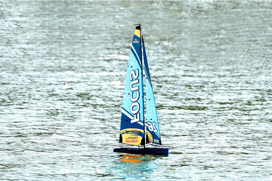 Joysway Focus V3 blue RC sailboat racing yacht gliding across open water with sunlight glinting on the surface.