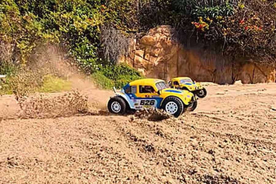Two yellow and blue Volkswagen Beetle RC buggies driving fast and kicking up dust on an outdoor dirt track.