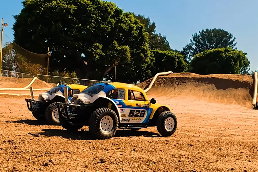 Two Tamiya Volkswagen Dune Beetle RC buggies parked on a dirt track outdoors with trees and a ramp in the background.