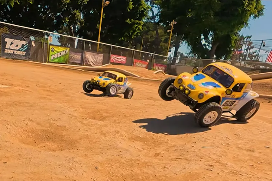 Two yellow and blue RC dune buggies racing on an outdoor dirt track with jumps and a safety fence background.
