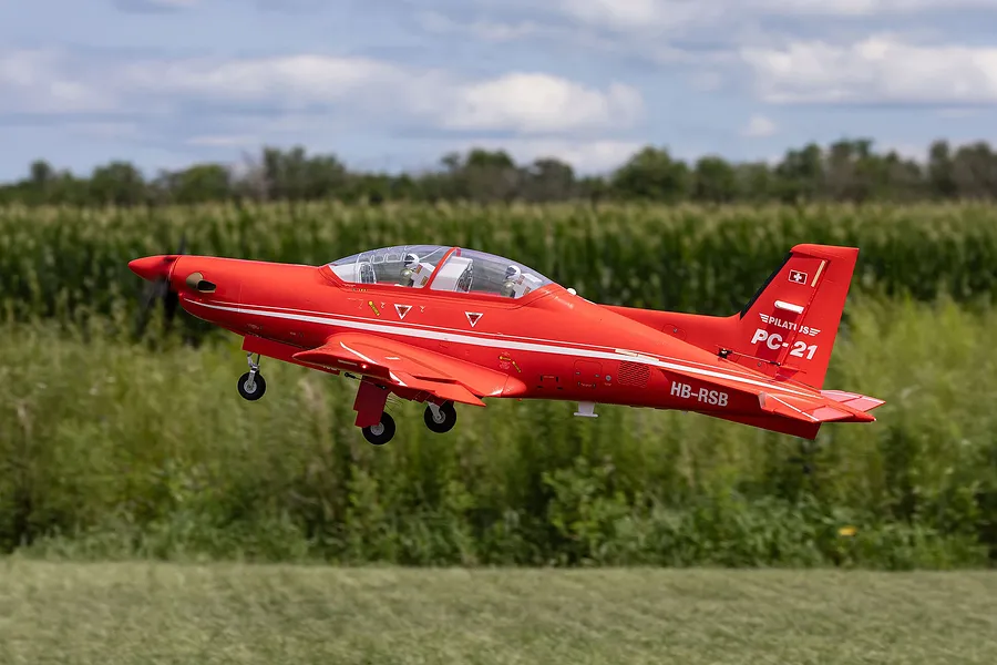 Red RC Pilatus PC-21 airplane in flight over a grassy field with landing gear extended.