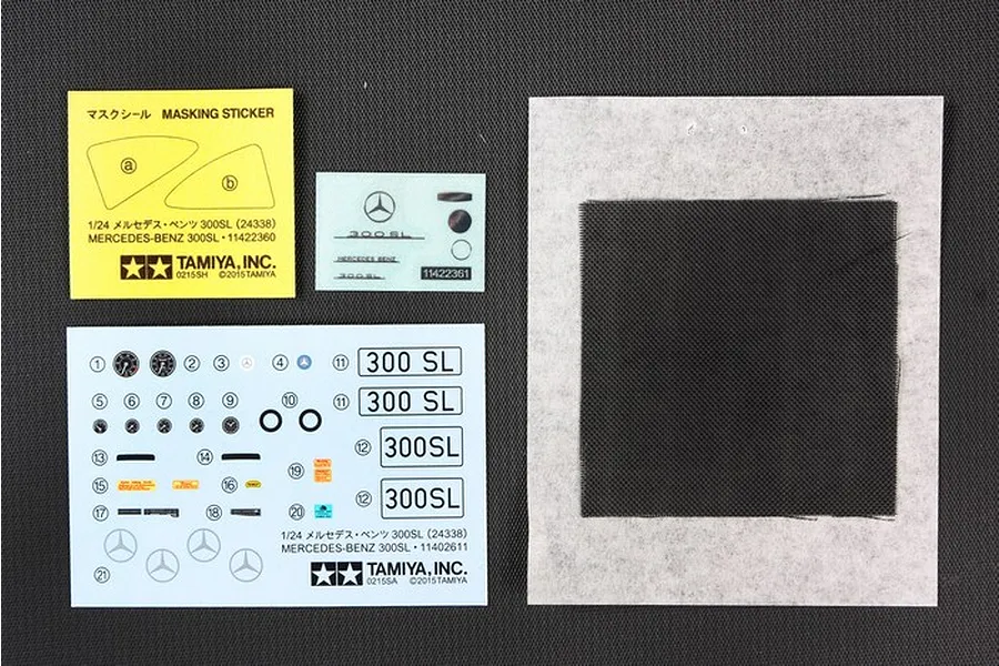 A set of Tamiya 1/24 scale Mercedes-Benz 300SL model kit decals, masking stickers, and a black mesh sheet on a dark textured background.
