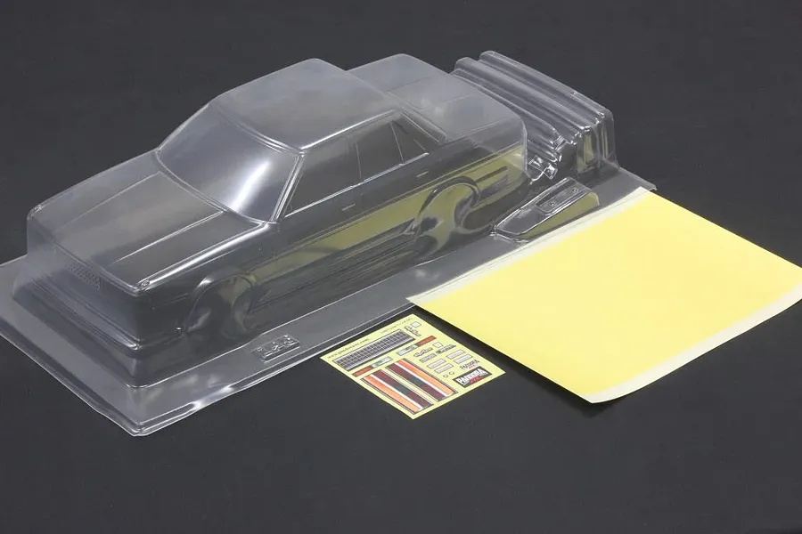Unpainted clear polycarbonate RC car body shell with a sticker sheet and masking sheet on a dark surface.