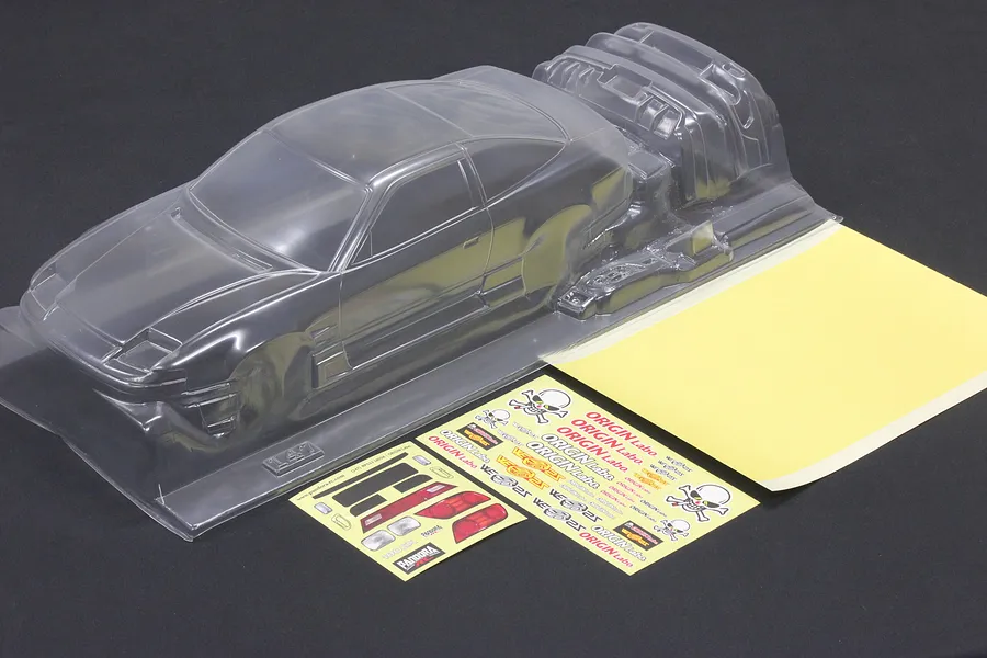 Unpainted clear RC car body shell with separate parts, decal sheets, and masking sheet on a dark background.