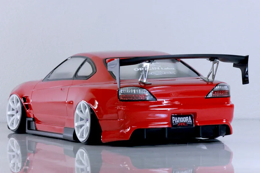 Rear three-quarter view of a red RC drift car with a large rear wing and white wheels on a reflective surface.