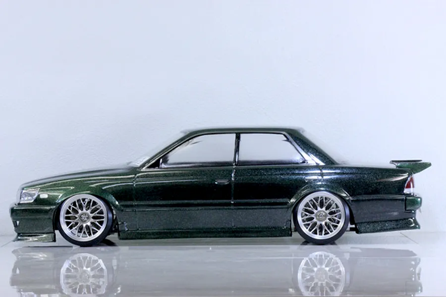 Side profile view of a painted green metallic RC drift car body shell mounted on a chassis with custom wheels against a white studio background.