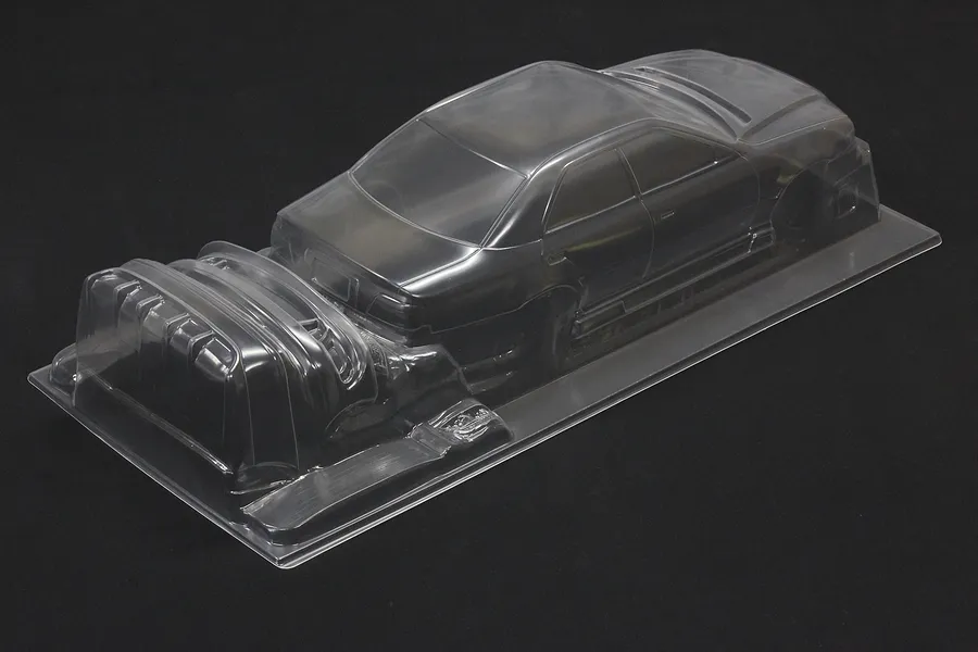Clear unpainted polycarbonate RC car body shell on a black background, showing sedan profile and separate front bumper parts.