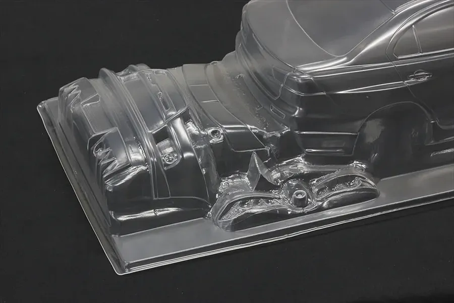 Unpainted clear polycarbonate RC car body shell showing rear section and molded bumper parts on a dark background.