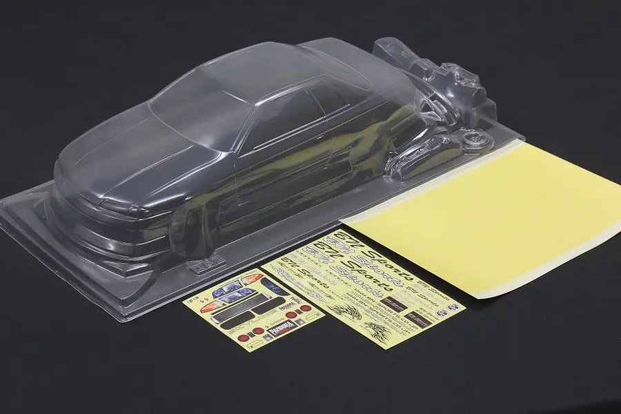 Unpainted clear polycarbonate RC body shell with a decal sheet and masking sheet on a dark surface.