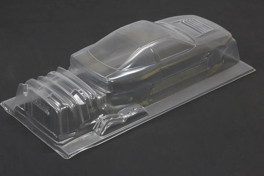 Clear unpainted polycarbonate RC car body shell on a dark surface, showing molded coupe and accessories.