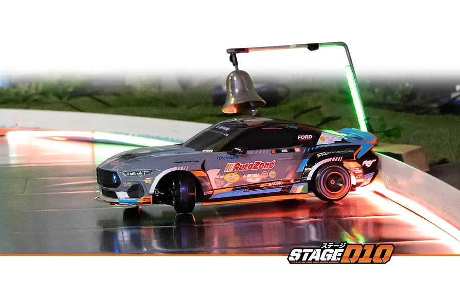 HPI Stage-D10 Ford Mustang drift RC car driving on an indoor track illuminated by red and green lights, passing under a suspended bell.