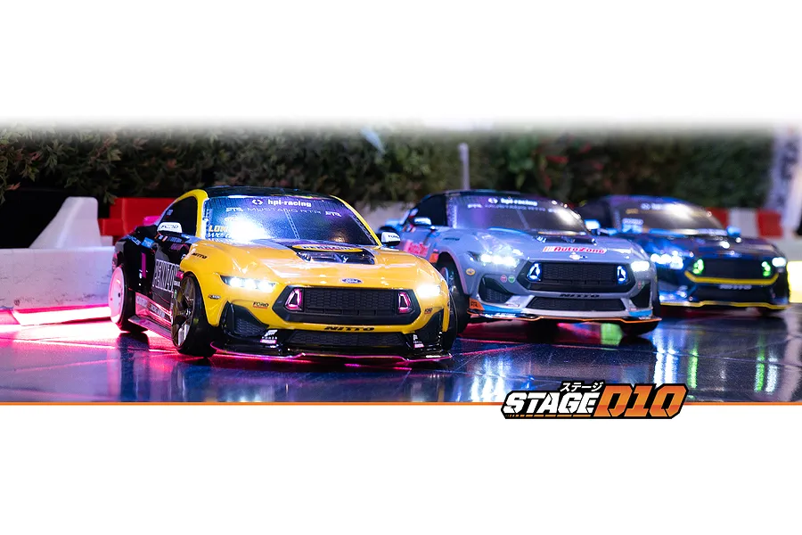 Three RC drift cars, including a yellow and black Mustang GT, driving on a reflective track with illuminated headlights.