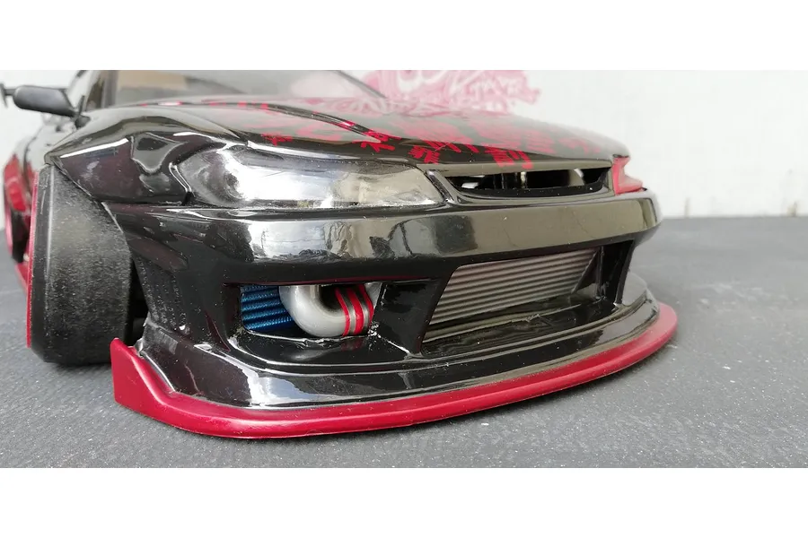 Close-up of a black RC drift car front bumper showing an installed aftermarket intercooler and piping behind the grille.