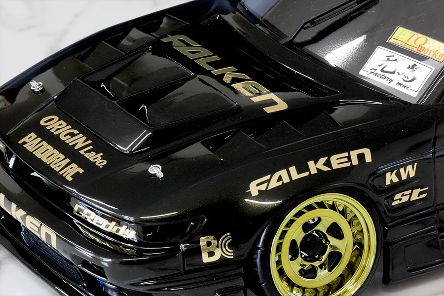 Close-up of a black RC drift car body with gold decals, custom vented hood, and gold rims on a light surface.