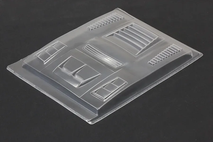 Unpainted clear polycarbonate RC body shell accessory sheet with various molded duct and vent parts on a dark surface.
